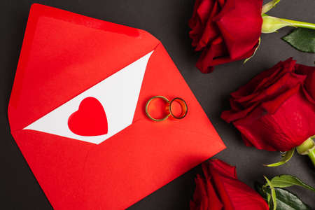 top view of red roses near envelope and golden rings on black