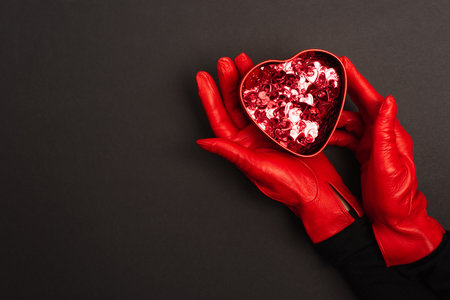 cropped view of woman in red gloves holding metallic heart-shaped box on blackの写真素材