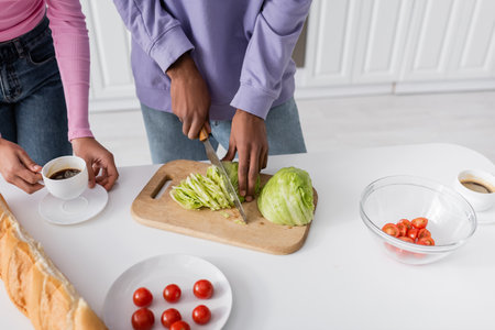 Cropped view of african american man cutting cabbage near girlfriend with coffee and baguette in kitchenの写真素材