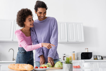 Positive african american woman pointing at cherry tomatoes while boyfriend cooking salad at homeの写真素材