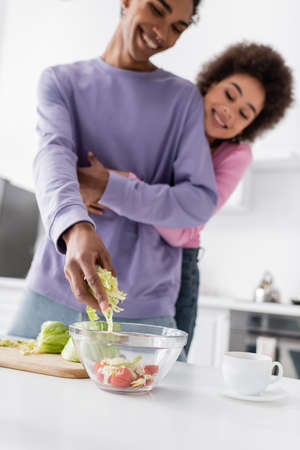 Blurred african american man cooking salad near cup and girlfriend at homeの写真素材