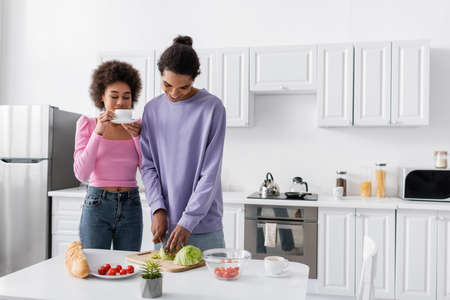 Smiling african american man cutting cabbage near girlfriend with coffee cup in kitchen