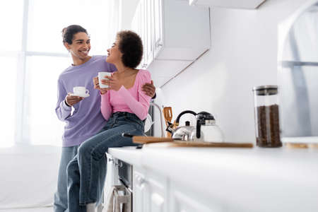 Young african american man hugging girlfriend with cup on worktop in kitchenの写真素材
