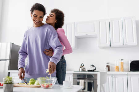 African american woman hugging and pouting lips near boyfriend cooking salad in kitchenの写真素材