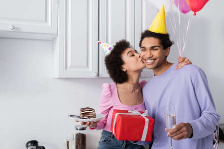 Positive african american woman holding cake and kissing boyfriend with gift and champagne at homeの写真素材