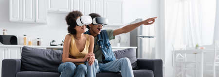 Excited african american man in vr headset pointing with finger near smiling girlfriend at home,...