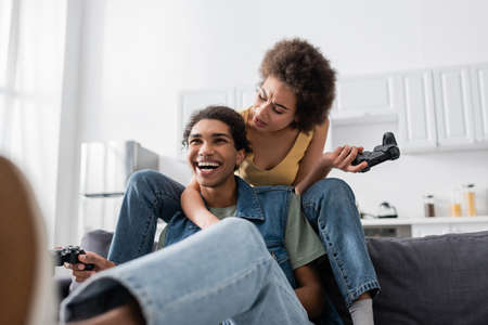 KYIV, UKRAINE - NOVEMBER 9, 2021: Displeased african american woman holding joystick near cheerful boyfriend on couchの写真素材