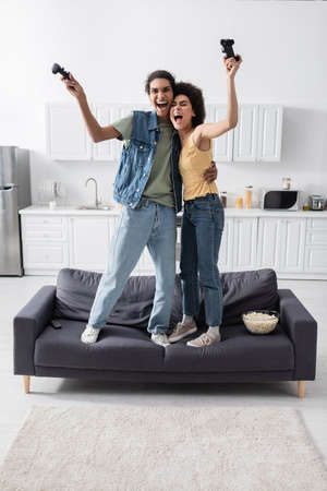 KYIV, UKRAINE - NOVEMBER 9, 2021: Excited african american couple holding joysticks on couch near...