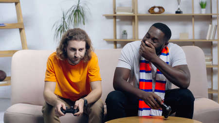 KYIV, UKRAINE - DECEMBER 22, 2021: upset man holding joystick near laughing african american friend covering mouth with handの写真素材