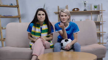 tense women sitting on couch with soccer ball and striped scarf and watching football match on tvの写真素材