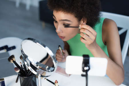 African american blogger applying mascara near cosmetic brushes and smartphone