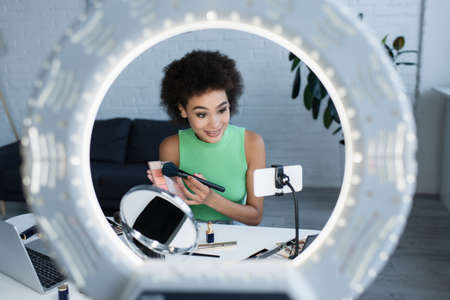 Young african american blogger holding blush near smartphone, mirror and ring light at home