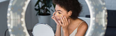 Young african american woman applying eye patches near mirror and blurred ring light, bannerの写真素材