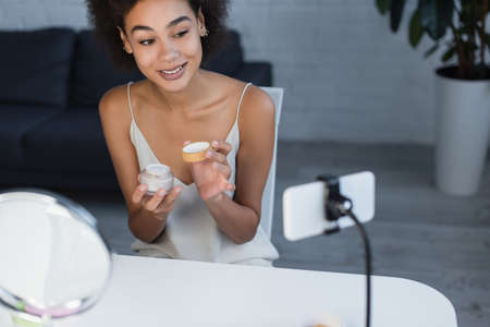 Cheerful african american blogger holding cosmetic cream near cellphone at homeの写真素材