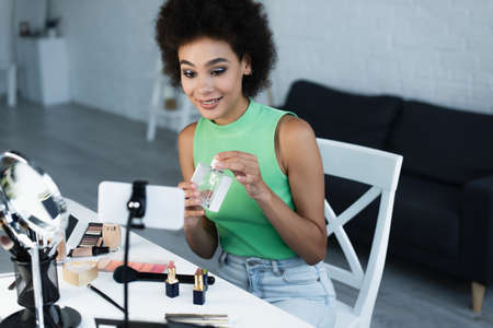 Cheerful african american blogger holding perfume near cosmetics and smartphoneの写真素材