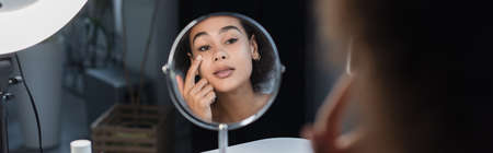 Young african american woman applying cosmetic cream near mirror and ring light at home, bannerの写真素材