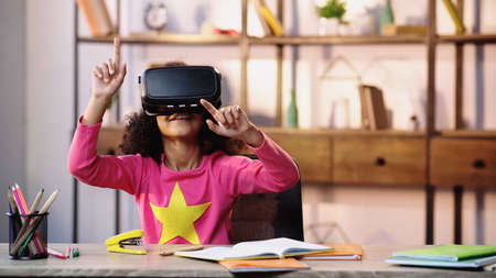 cheerful african american child in vr headset gesturing while sitting at deskの写真素材