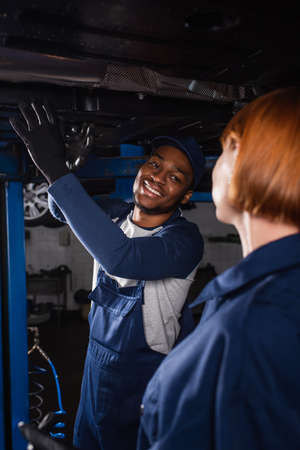 Smiling african american mechanic looking at blurred colleague near auto in serviceの写真素材