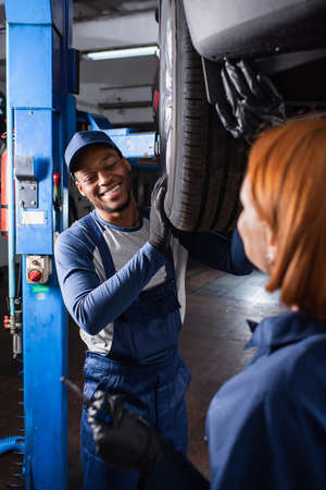 Positive african american mechanic working with car wheel and blurred repairwoman in serviceの写真素材