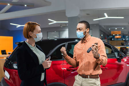 excited african american man in medical mask holding key and showing win gesture near car and sellerの写真素材
