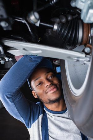 african american repairman smiling at camera while inspecting shock absorber of carの写真素材