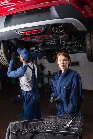Redhead mechanic holding tool and looking at camera near car and african american colleague in garageの写真素材