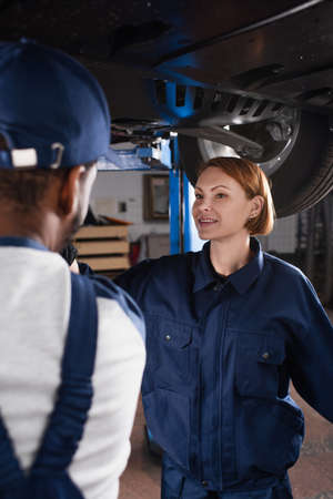 Smiling mechanic in uniform talking to blurred african american colleague under car in garageの写真素材