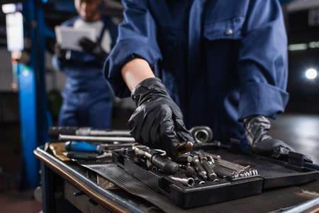 Cropped view of mechanic in gloves taking tool near blurred colleague in car service