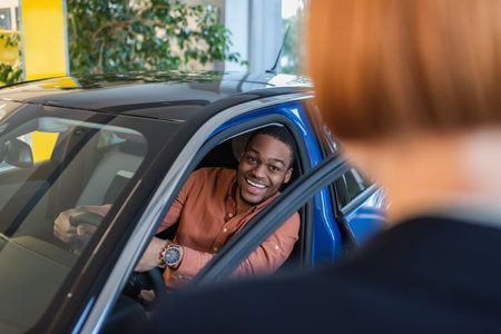 cheerful african american man looking at blurred car dealer while sitting in carの写真素材