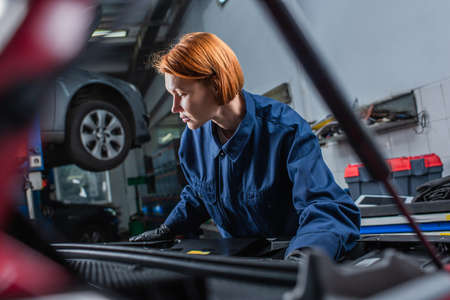 forewoman in uniform inspecting blurred car with open hood in workshopの写真素材