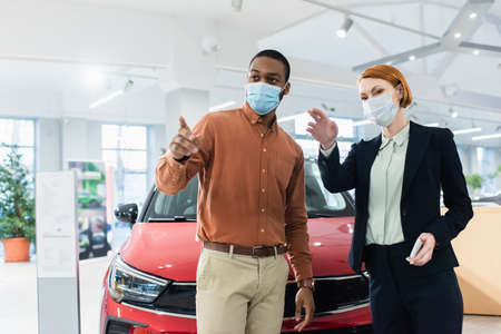 car dealer and african american client in medical masks pointing with fingers in car showroomの写真素材
