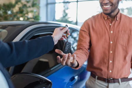 cropped view of car dealer giving key to african american client smiling at blurred background