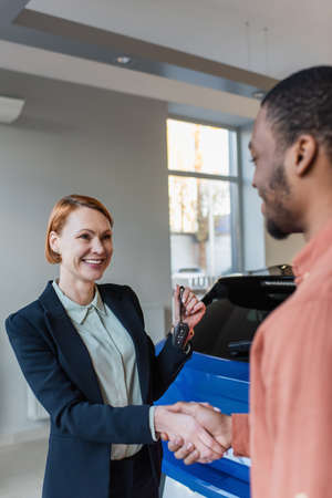 happy car dealer holding key and shaking hands with blurred african american customerの写真素材