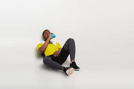 full length of african american sportsman drinking water from sports bottle while sitting on grey