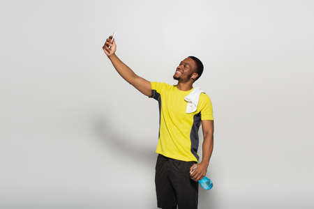 happy african american sportsman with earphone in ear taking selfie on grey