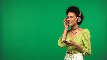 happy african american woman in wireless headphones holding smartphone isolated on green