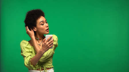 african american woman in blouse looking at smartphone and adjusting hair isolated on greenの写真素材