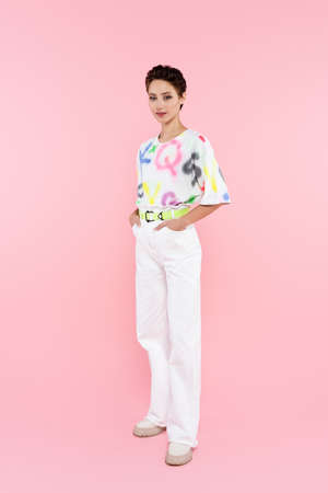 full length view of stylish smiling woman standing with hands in pockets of white trousers on pinkの写真素材