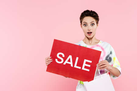thrilled woman with shopping bag and red card with sale lettering looking at camera isolated on pink
