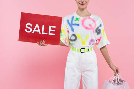 partial view of smiling woman holding shopping bags and red card with sale lettering isolated on pinkの写真素材