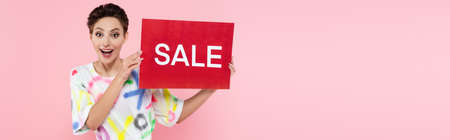 astonished brunette woman showing credit card with sale lettering isolated on pink, bannerの写真素材