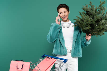 smiling woman with small fir tree talking on smartphone near cart with shopping bags isolated on greenの写真素材