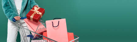 partial view of trendy woman with present near shopping cart with purchases isolated on green, bannerの写真素材