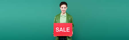 stylish brunette woman holding placard with sale lettering while smiling at camera isolated on green, bannerの写真素材