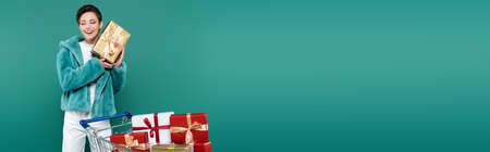 pleased woman with closed eyes holding gift box near shopping cart with presents isolated on green,...