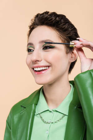pleased brunette woman with short hair applying mascara isolated on beigeの写真素材