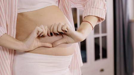 Cropped view of pregnant woman showing heart sign near belly at homeの写真素材