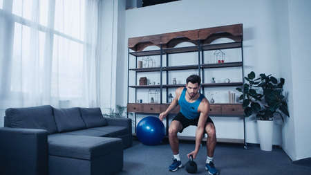 bearded young sportsman in blue tank top exercising with kettlebell at homeの写真素材