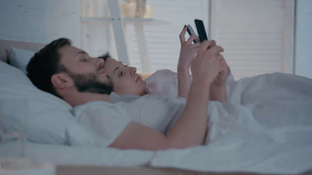 Young woman using smartphone near husband on bed at homeの写真素材