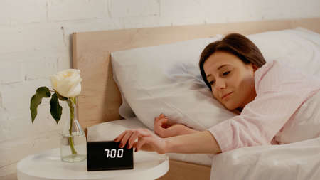 Young woman turning off clock near flower in bedroom